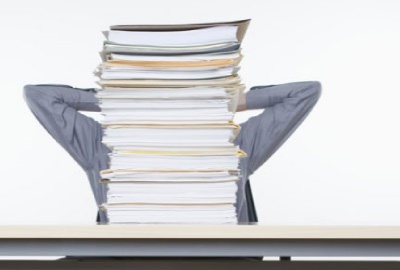 Image of person sitting behind a towering stack of papers punctuating the need for project document management.